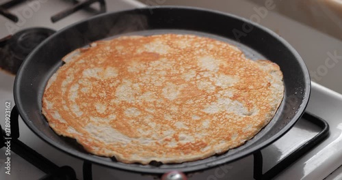A golden-brown pancake cooking in a black frying pan on a gas stove. The pancake has a smooth texture and is evenly cooked.