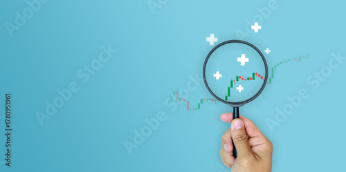  Financial growth, investment analysis, profit growth, and positive economic outlook assessment. Hand holding magnifying glass to focus on bullish candlestick chart with plus sign on blue background.