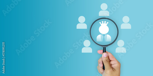 Принти на полотні Hand holding magnifying glass focusing on a crowned figure among people icons on blue background, symbolizing leadership, recruitment, human resources management, and finding top performer potential
