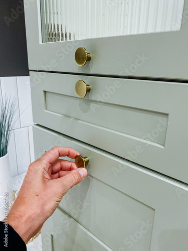 Hand adjusting brass cabinet knob over mint drawer, gentle grip suggesting human interaction, warm bathroom tile backdrop, closeup detail conveying tactile lifestyle moment and modern