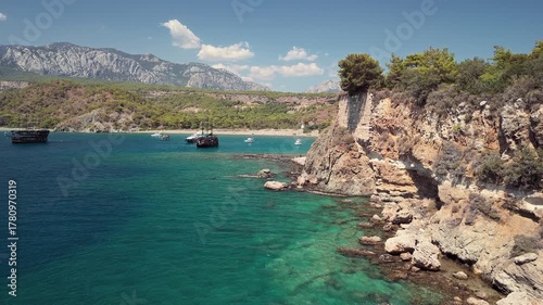 Aerial view of Phaselis Bay with sailing ships and Mount Olympos in the background. Clear blue water and scenic Mediterranean coastline near Antalya, Turkey. Captures scenic coastal beauty in Turkey.