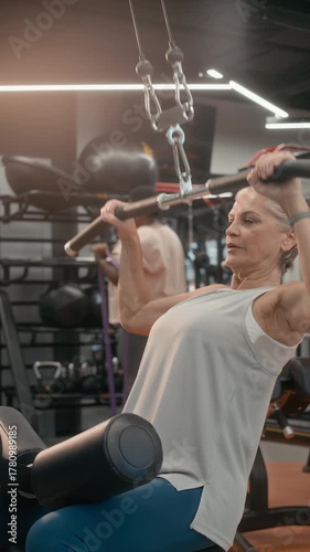 Vertical slowmo shot of athletic mature Caucasian woman in activewear working out in modern gym using lat pulldown machine