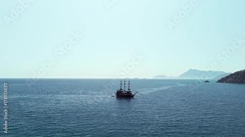 Aerial view of a sailing ship with tall masts cruising through the blue waters of the Gulf of Antalya, Turkey. Clear sky, calm sea, and scenic coastal mountains in the distance.