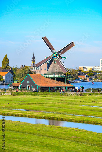 A traditional windmill and historic wooden houses on the waterfront of a lake or wide river, with green fields in the foreground.