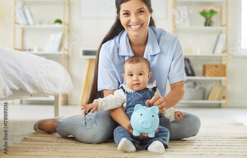 Canvastavla A smiling mother sits on the floor with her baby in a bright nursery, holding a blue piggybank to teach early savings and parental financial skills