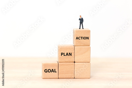 Wooden cubes arranged as ascending steps labeled ACTION, PLAN, and GOAL with a miniature businessman figure standing on the top block. Concept of business success, achievement, and strategic planning