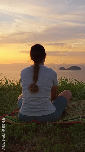Vertical video. Woman Sitting Cross-Legged on Mat Looking at Sea and Islands during Sunset, Rear View, Wide Angle, Tranquility and Self Reflection Concept