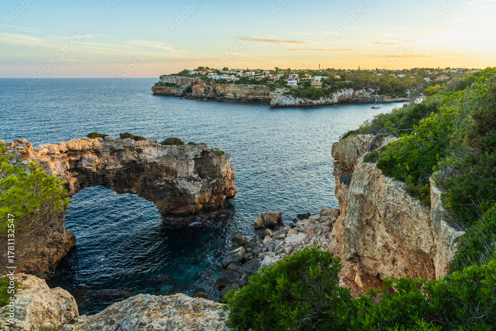 Obraz premium Majestic stone arch in sea at Cala Santany?, Mallorca, Spain during sunset. Serene seascape with glowing sky and coastal rock formations.