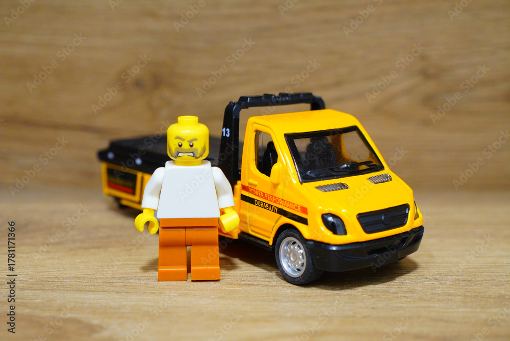 Obraz premium Lego figure stands next to a yellow toy tow truck on a wooden surface