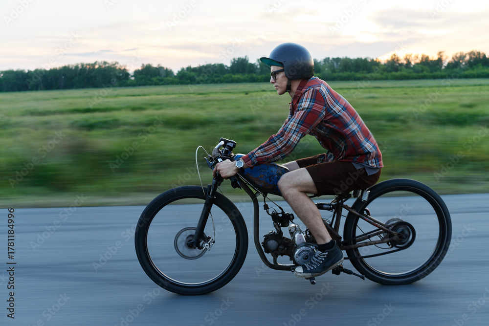 Obraz premium Side view portrait of young hipster man riding retro bike on deserted countryside road at dusk