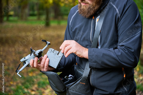 Canvas Print A man with a bushy beard prepares his drone for flight in a peaceful park filled with trees