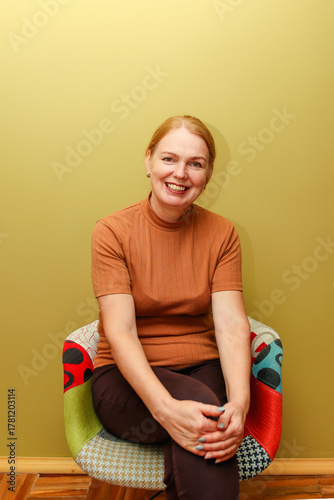 Smiling middle-aged woman with red hair in brown top sits on colorful patchwork chair against light green wall. portrait of a middle-aged woman 45 years old, with a warm smiling expression