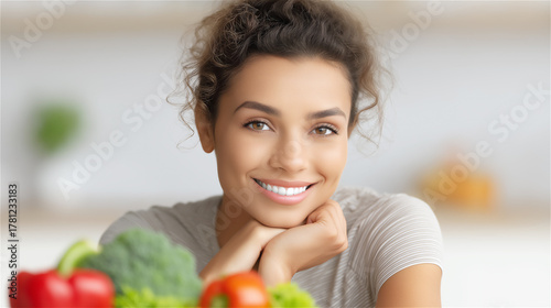 Young woman smiling in kitchen with fresh vegetables on table, concept of healthy eating and balanced lifestyle.