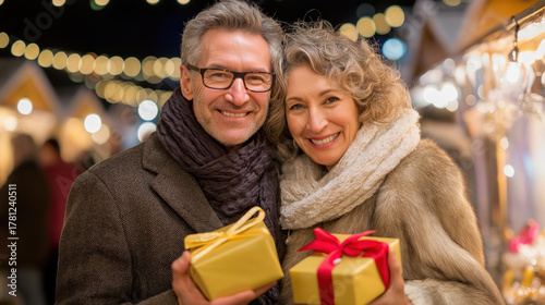 Smiling mature couple holding gifts at a Christmas market at night. Happy senior man and woman enjoying a festive holiday evening together
