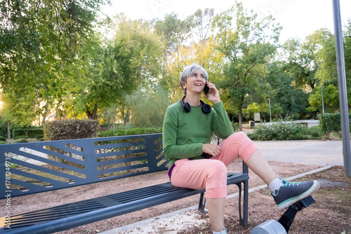 Cheerful middle-aged woman with gray hair sitting comfortably on a park bench, talking on her mobile phone and enjoying the calm afternoon outdoors.