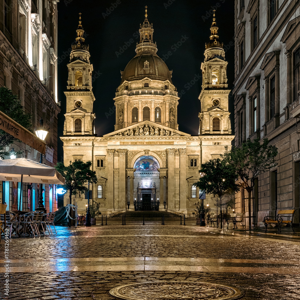 Fototapeta premium St. Stephen’s Basilica at night illuminated by streetlights and reflections on wet cobblestone street in the historic center of Budapest, Hungary