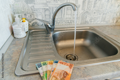 Belarusian rubles at an open water tap in the kitchen, water savings, and increased utility bills