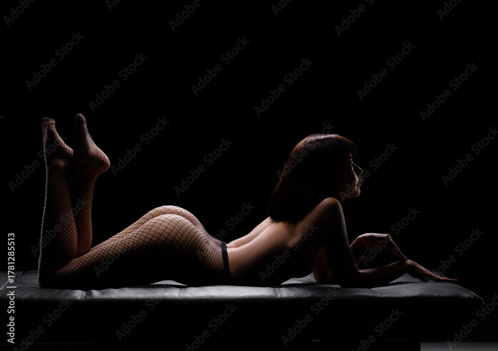 Obraz premium Artistic nude portrait of a woman in fishnet tights lying on a couch under dramatic backlight. Minimalist style, play of light and shadow, elegant body curves and sensual mood.