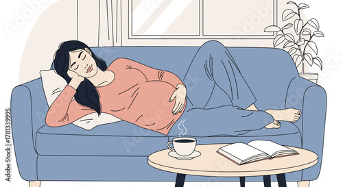 Young asian pregnant female relaxing on a blue sofa, holding her belly, with a book and steaming cup of coffee on a table nearby.