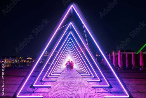 Purple coloured gate of light or purple light tunnel installation made of triangular neon lights at night as blurred people walk through the bright deep passage