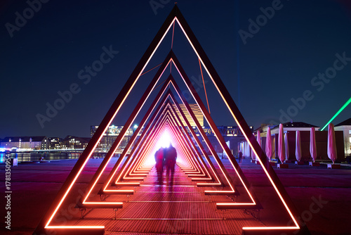 Blurred people like aliens cross a light tunnel with visible stars. Light tunnel or gate of light installation consists of many triangular gates lit by led bright lights