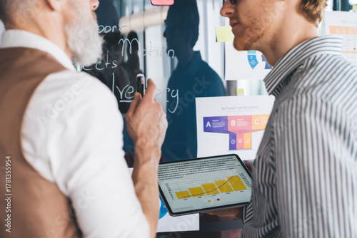 Senior executive points at wall while young professional holds tablet. Scene highlights collaborative analytics, visual planning, and digital tools in real-time team communication.