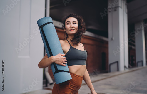 Athletic young woman smiling while carrying yoga mat over shoulder. Conceptual lifestyle photo symbolizing empowerment, mindfulness, wellness, and connected digital generation identity.