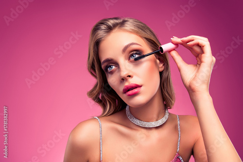 Young stylish woman applying mascara on eyelashes in a glamorous pink background, showcasing beauty