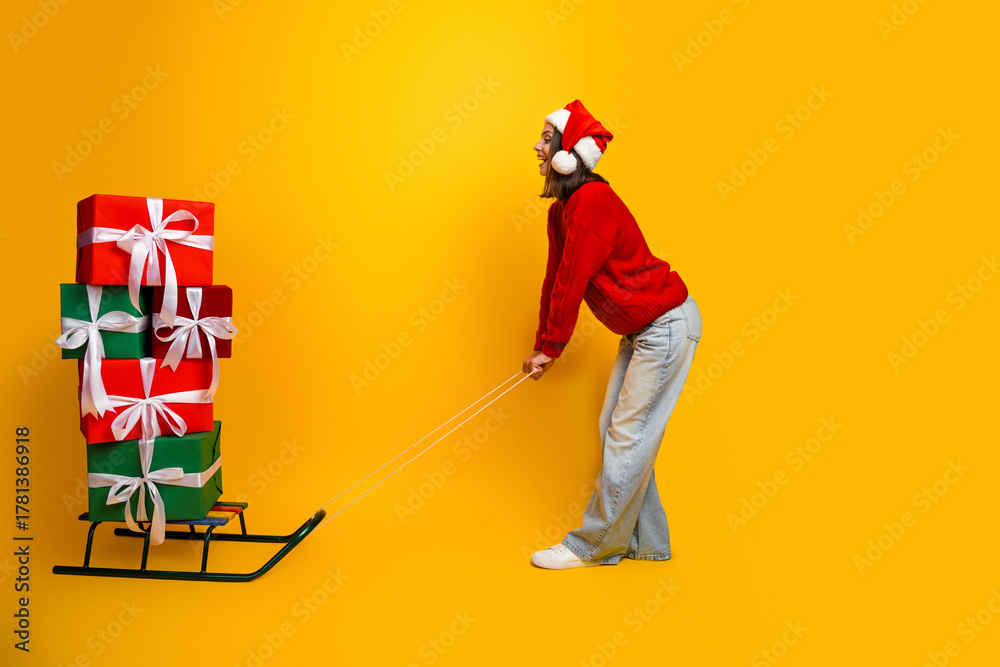 Naklejka premium Funny Santa helper pulls stacked gifts on a sleigh in a bright yellow studio wearing red sweater and hat for holiday themed stock image