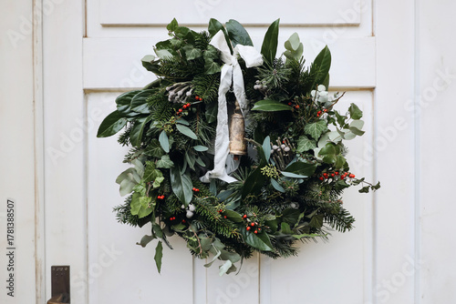 Beautiful Christmas wreath with greenery, eucalyptus, holly and red berries hanging on white door. Rustic ribbon, vintage bell. Natural festive floral decoration for winter holiday season, advent.