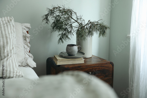 Winter bedroom. Cup of coffee tea on books, wooden bedside table. Pine tree branches in vase. Sage green wall. Neutral linen pillows frills. Blurred blanket foreground. Christmas interior still life.