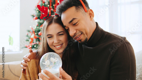 Photos A joyful multiracial couple embraces in a warm setting, admiring a snow globe while celebrating Christmas next to a decorated tree, filled with holiday spirit