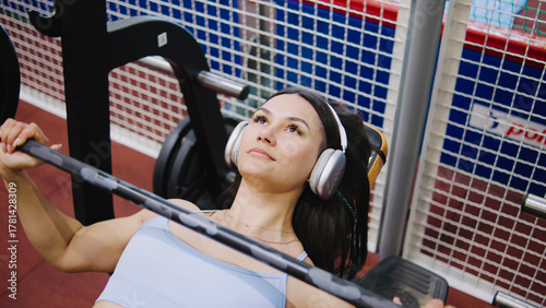 A woman exercises on a bench press in a gym. She is wearing headphones and concentrating on her workout while surrounded by gym equipment during the day.
