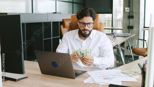 A man in casual attire counts cash while sitting at a stylish office desk, focused on his laptop in a well-lit workspace during the day.