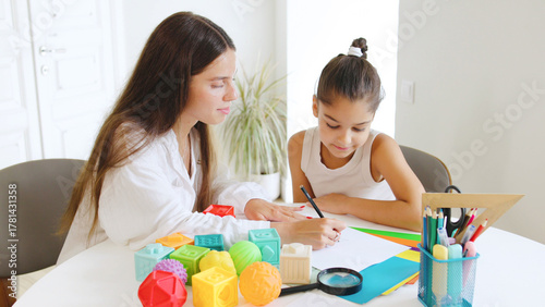 A speech therapist guides a young girl in a productive session, actively engaging her with colorful educational materials on a round table in a well-lit room.