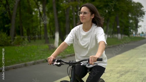 Young woman in white t shirt and black leggings riding bicycle in park front view. 4k