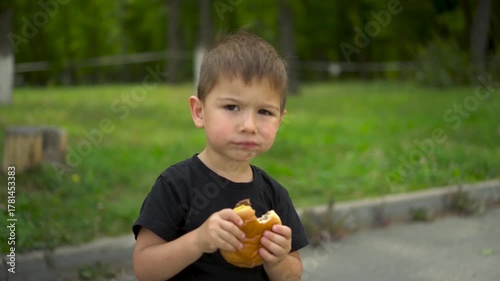 Little boy eats cheeseburger in park and looks at camera portrait shot 4k
