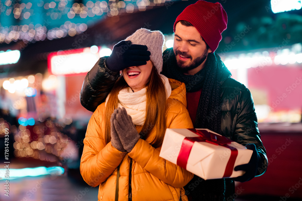 Obraz premium playful couple share a surprise gift under bright christmas lights in a festive city market night scene