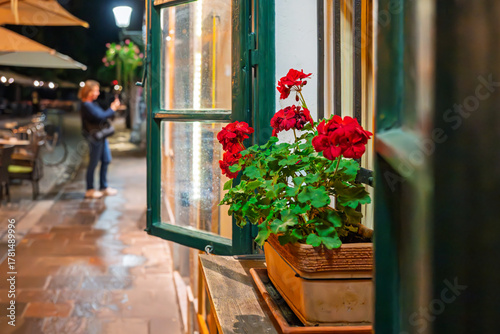 Red geraniums in a flower box sit on an open window sill near a sidewalk cafe on the Ljubljanica River embankment at night, in the historic old town of Ljubljana, Slovenia.