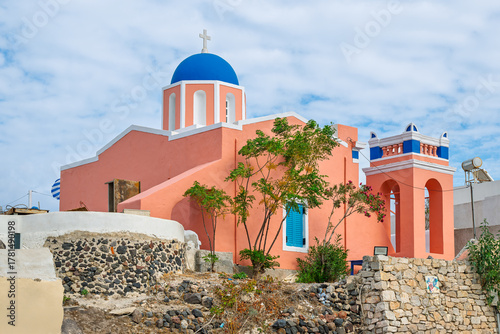 Fototapeta A vibrant salmon pink colored blue dome church in the hillside village of Oia, Greece, on the island of Santorini