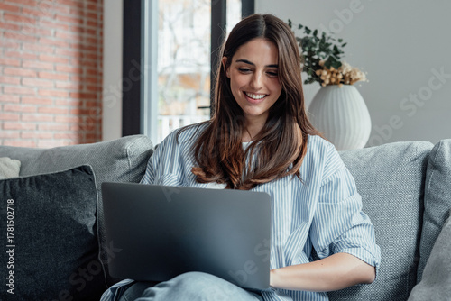 Concentrated millennial girl sit on couch working on laptop browsing internet at home during lazy weekend, focused young woman freelancer busy using computer surfing wireless web shopping online