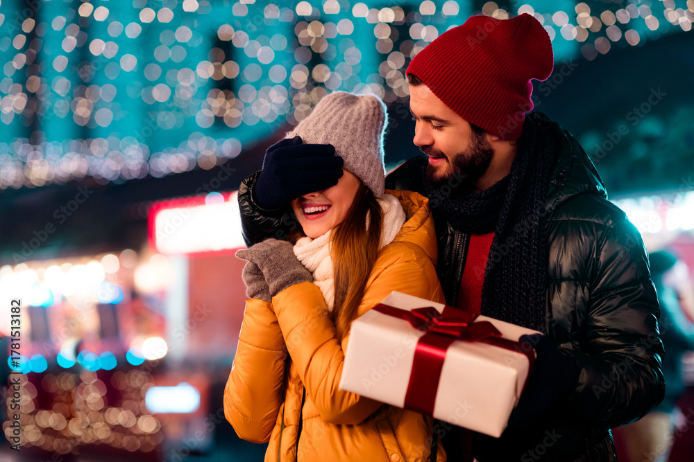 Obraz premium Joyful couple at a festive city market share laughter while gifting a wrapped present under sparkling lights