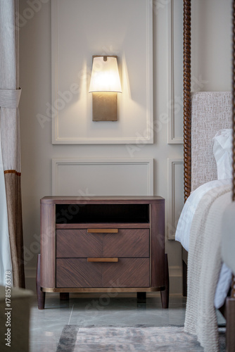 Luxury bedroom interior. Dark wood nightstand with brass handles, by a four-poster bed. A modern wall sconce lights a beige wall with molding.