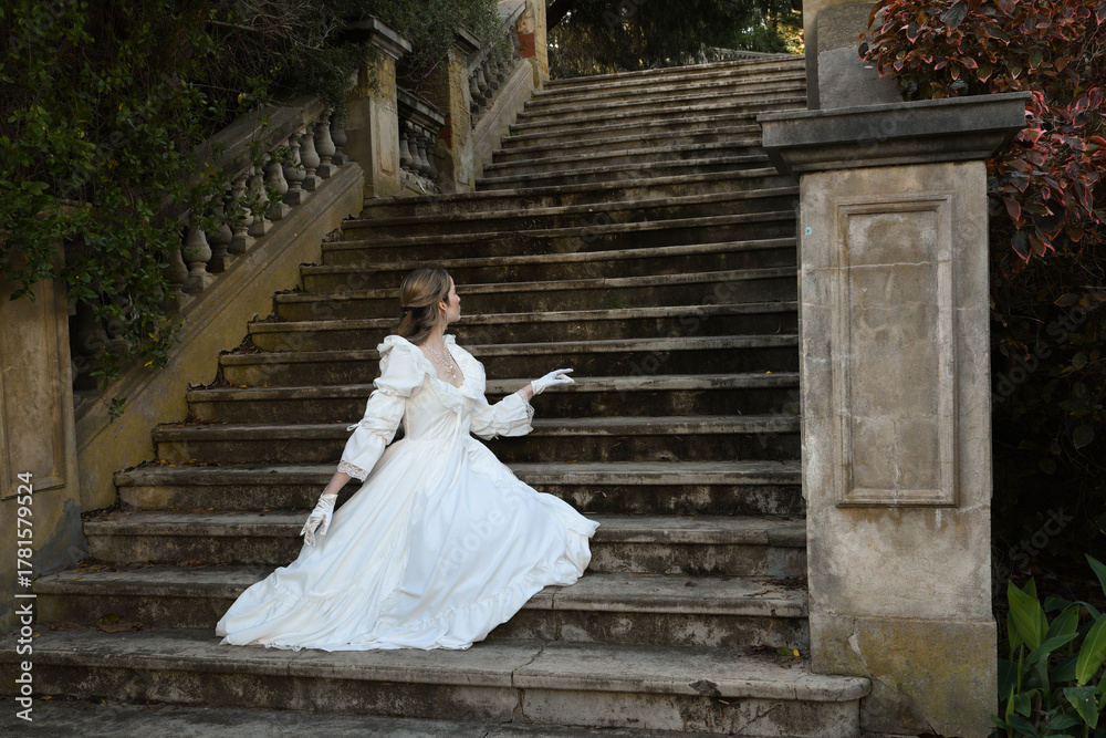 Obraz premium back view figure of female blonde model wearing white fantasy wedding gown. romantic sitting pose looking away on staircase of historical fairytale castle location