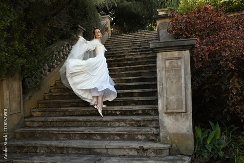 portrait of beautiful young blonde model wearing historical white wedding gown and fantasy cloak. wandering ethereal figure walking away on historical staircase of fairytale castle location.