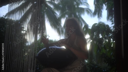  A girl in a white dress plays a handpan in a lush tropical setting, with palms.