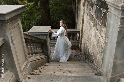 full length portrait of beautiful blonde female model wearing historical white gothic wedding gown, walking away back view, down staircase of romantic fairytale castle location with stone balconies