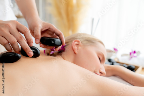 Hot stone massage at spa salon in luxury resort with day light serenity ambient, blissful woman customer enjoying spa basalt stone massage glide over body with soothing warmth. Quiescent