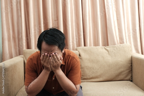 Upset Southeast Asian man sitting on a beige sofa with his face buried in his hands, expressing stress or emotional distress.
