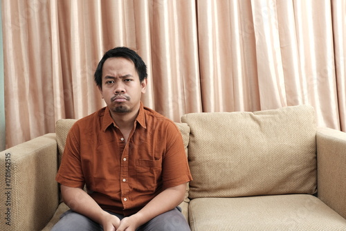 Southeast Asian man in casual clothing sitting on a beige sofa with a frustrated facial expression, appearing upset or disappointed.
